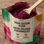 A stand-up pouch of Tiora 100% natural custom blend powder with a wooden spoon resting inside, set on a rustic wooden surface. The pouch has a white central label with black text and colorful abstract floral artwork in pink, green, teal, and yellow.