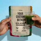 A person holds a stand-up pouch labeled Tiora Your Custom Blend in front of a light blue background, with a hand holding a clear glass of green drink to the right. The pouch has a cream center with black text and multicolored floral-style borders at the sides.